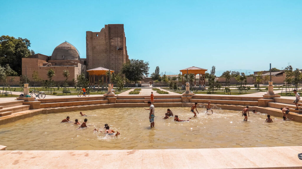 Shahrisabz in the middle of a redevelopment plan back in 2015. In the photo, a fountain with water and many children bathing in it. In the background, the Dorus Saodat complex.  